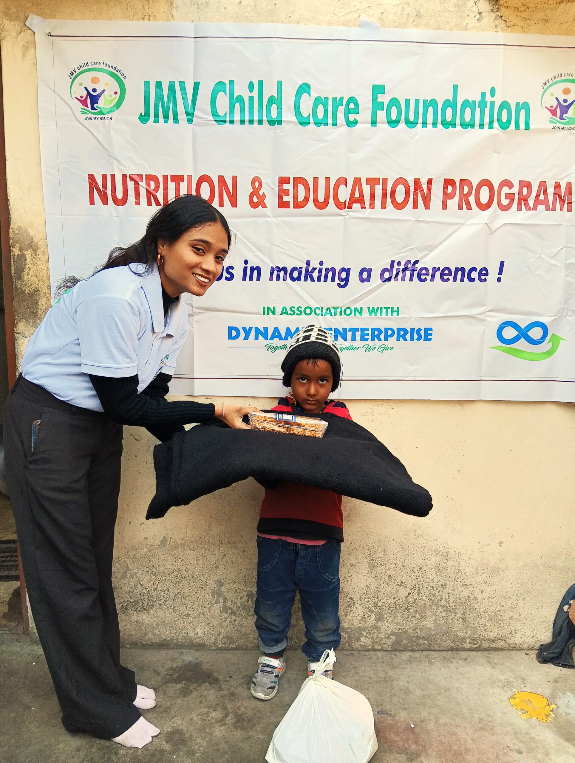 Supporter handing a clothes and sweet box to a visually impaired kid, both appearing happy.”
