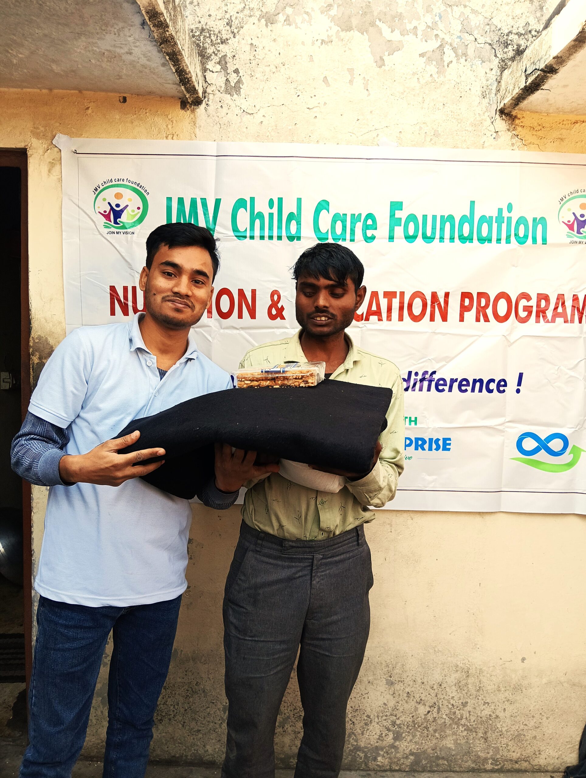 Supporter giving clothes and sweet box to a visually impaired boy, both appearing happy.”
